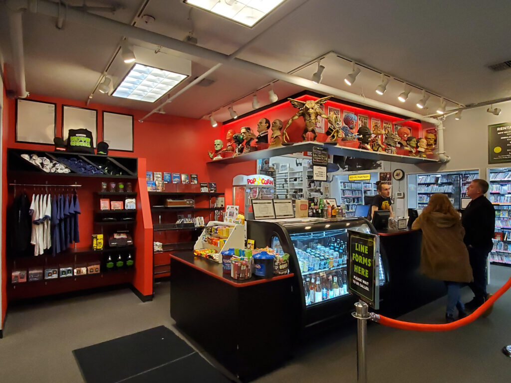 Concession stand at Movie Madness Miniplex with snacks, drinks, movie memorabilia, and two customers interacting with the attendant.
